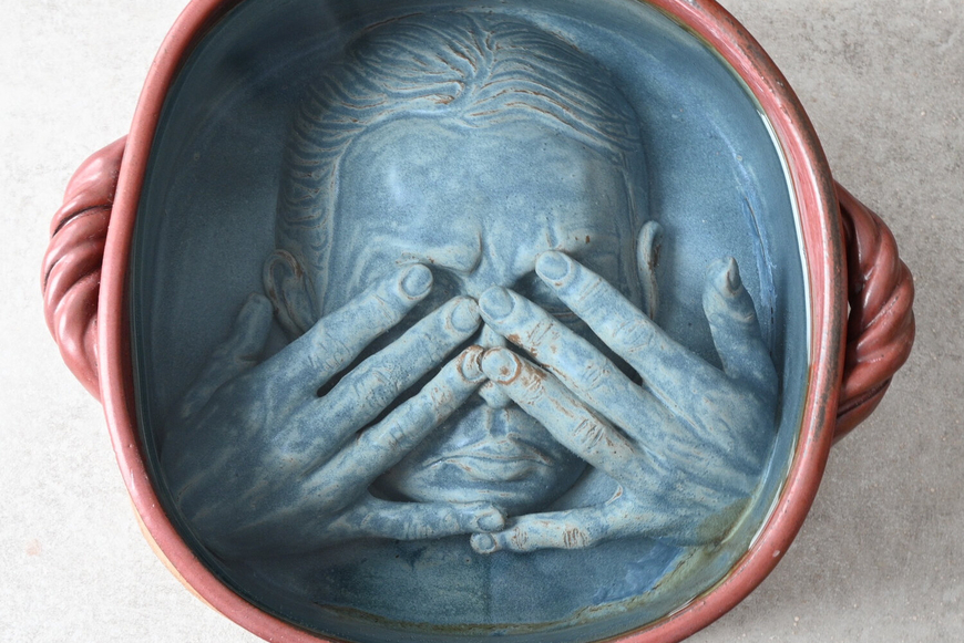 Ceramic vessel with red edges and a blue bottom. In the blue base, hands cover the eyes of a face contorted in anguish. 
