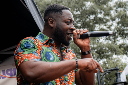 Edwin Owusu wearing a red green and blue shirt sings into a microphone outdoors.