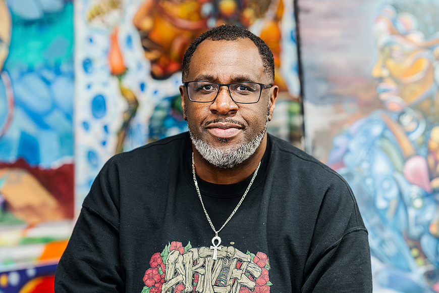 Will Kasso Condry of Juniper Creative Arts wears a black shirt and a chain with an ankh and sits in front of several colorful portrait paintings