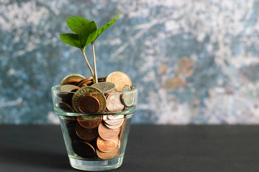 glass tumbler filled with coins with a plant growing out of it. Sitting on a dark gray surface with variegate gray-white-blue-brown background.