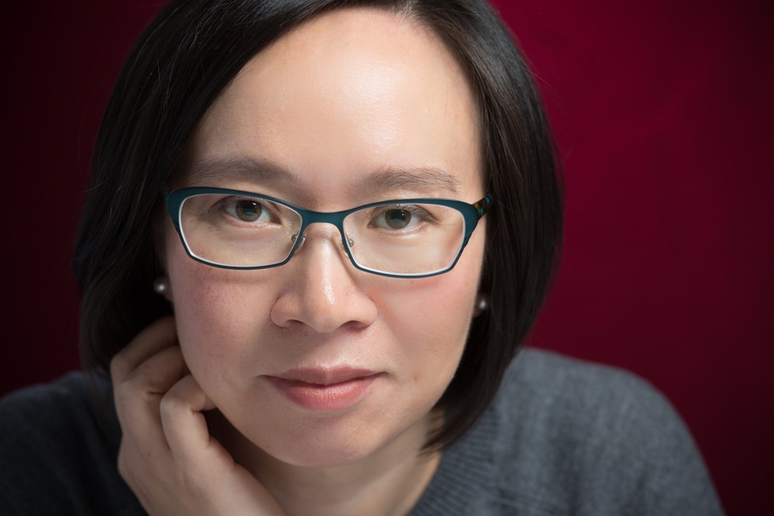 headshot of author Malinda Lo. Her expression is serious but approachable and she wears glasses with midnight blue frame, small earrings, and a grey sweater, all on a burgundy background.