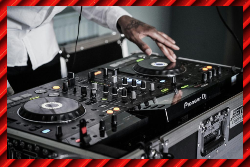 Color photo shows a DJ's left hand on a turntable and mixer all-in-one. Photo is set against a background of rich red diagonal striping.