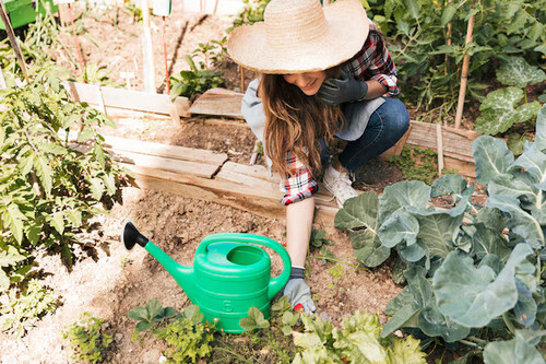 A woman squats in a garden next to a green watering can and has her hands in the soil working with plants