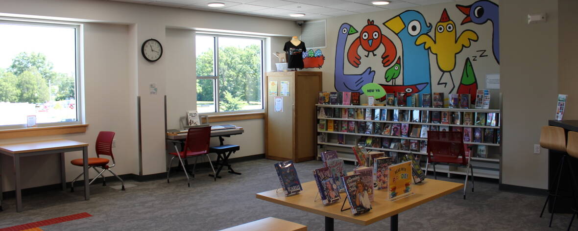 Two windows let in daylight over a piano, tables and chairs for work, a table with several books on display, a colorful mural of cartoony birds, and shelves of new YA books.