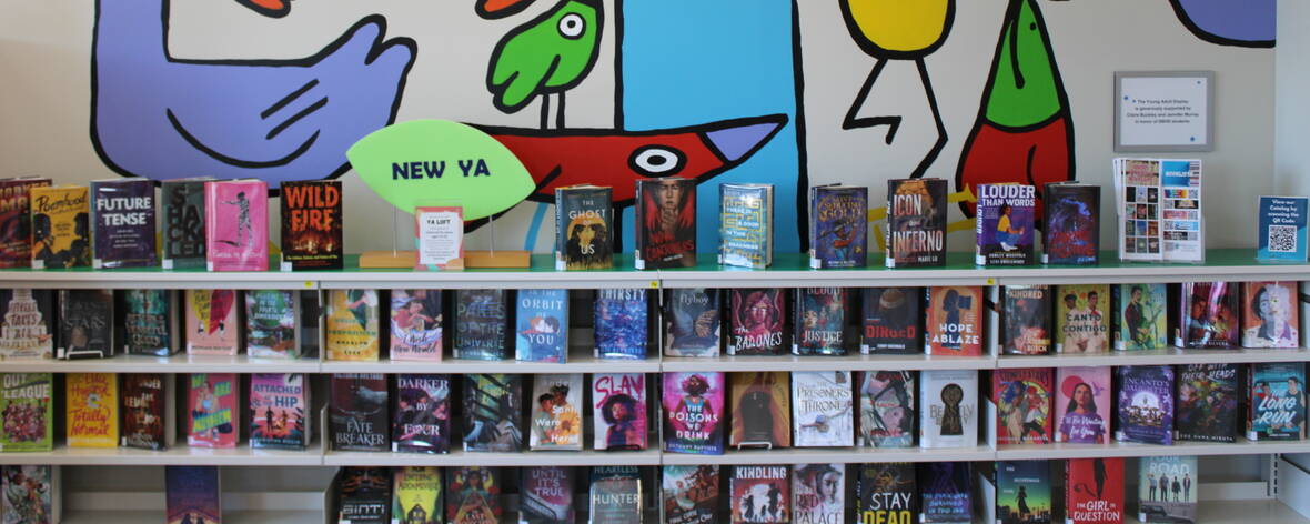 A close up of a colorful mural of cartoonish birds, a green sign says New YA and there are four shelves of a variety of new YA books on display