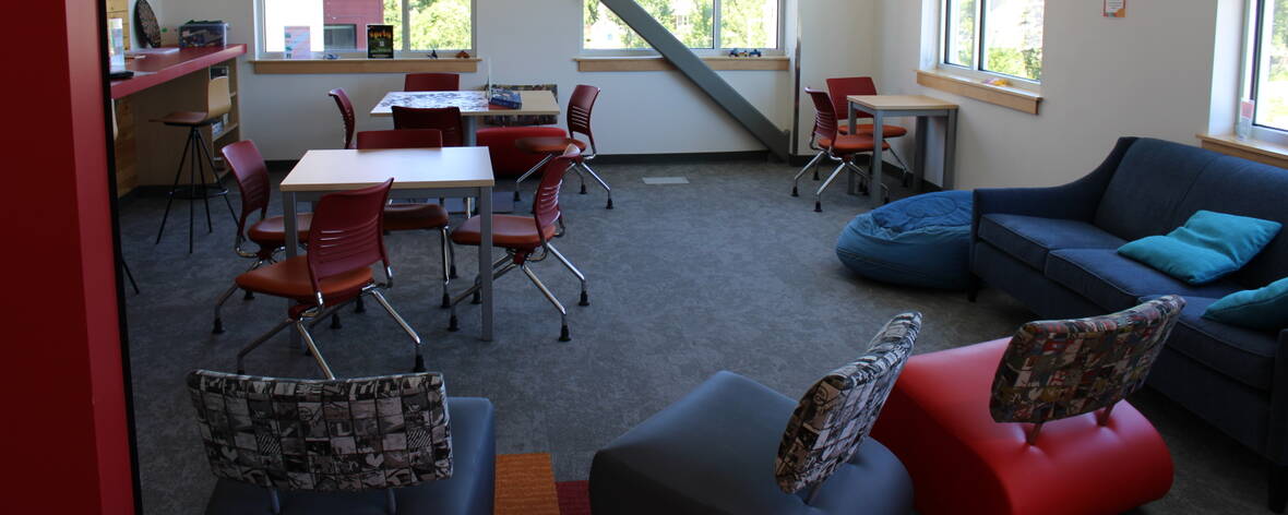 Sunlight pours in from four windows over blue and orange couches and chairs are placed around for seating along with tables for doing work or games