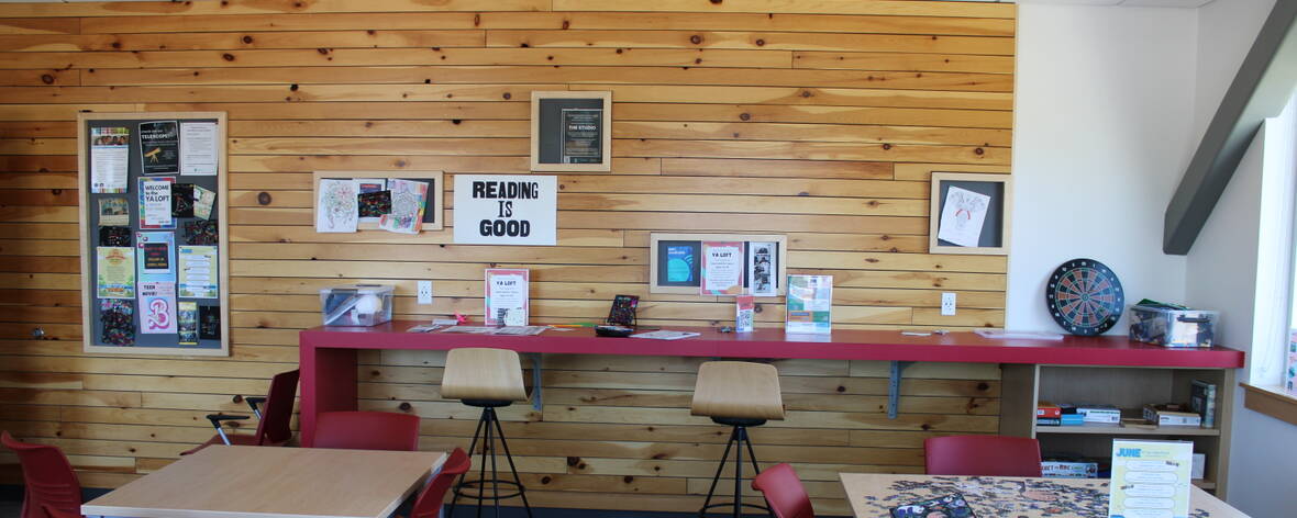 A wood paneled wall has signs, posters, and a community board. Two chairs sit at a long red desk with activities for teens. A puzzle sits unfinished on a table in the foreground.