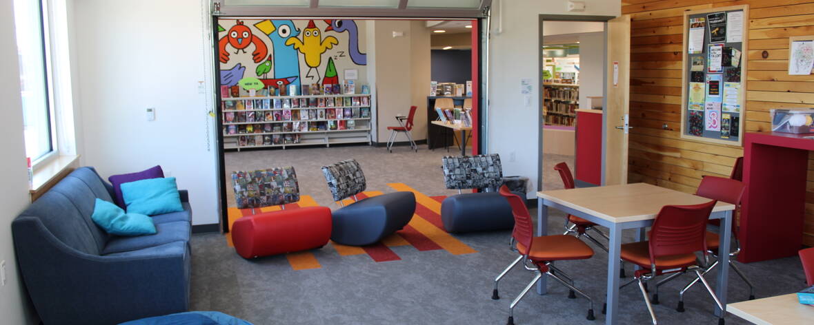 A vantage point from the corner of the Teen Loft. Blue and orange couches and chairs are placed around for seating along with tables for doing work or games. Beyond the seating is a mural of colorful birds with new YA books on the shelves below it.