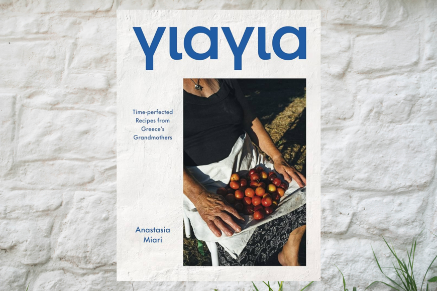 A photograph of a pile of red, orange, and yellow fruits held in a tea towel on the lap of a person wearing a skirt. Strong sunlight is shining on her. Book title Yiayia and cover text are in blue type.