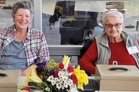 Two women sitting at a table with flowers collecting money from the book sale.