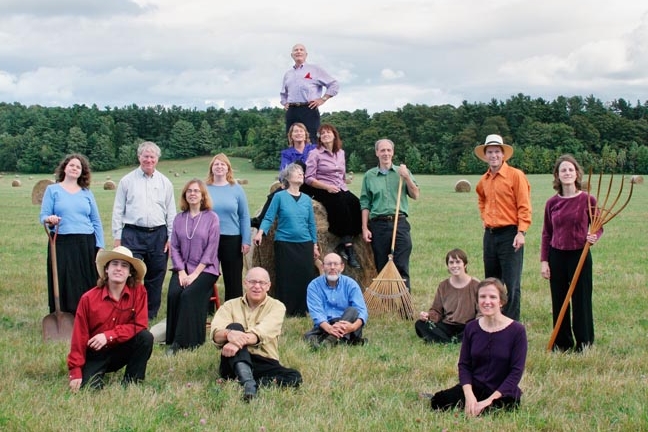 Group of 16 people sitting and standing in a green field with a variety of farm tools, wearing colorful solid shirts.