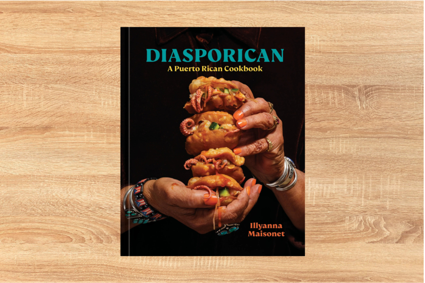 A light wood background with the cover of Diasporican: A Puerto Rican Cookbook. Two hands with orange painted nails hold a dish from the cookbook.